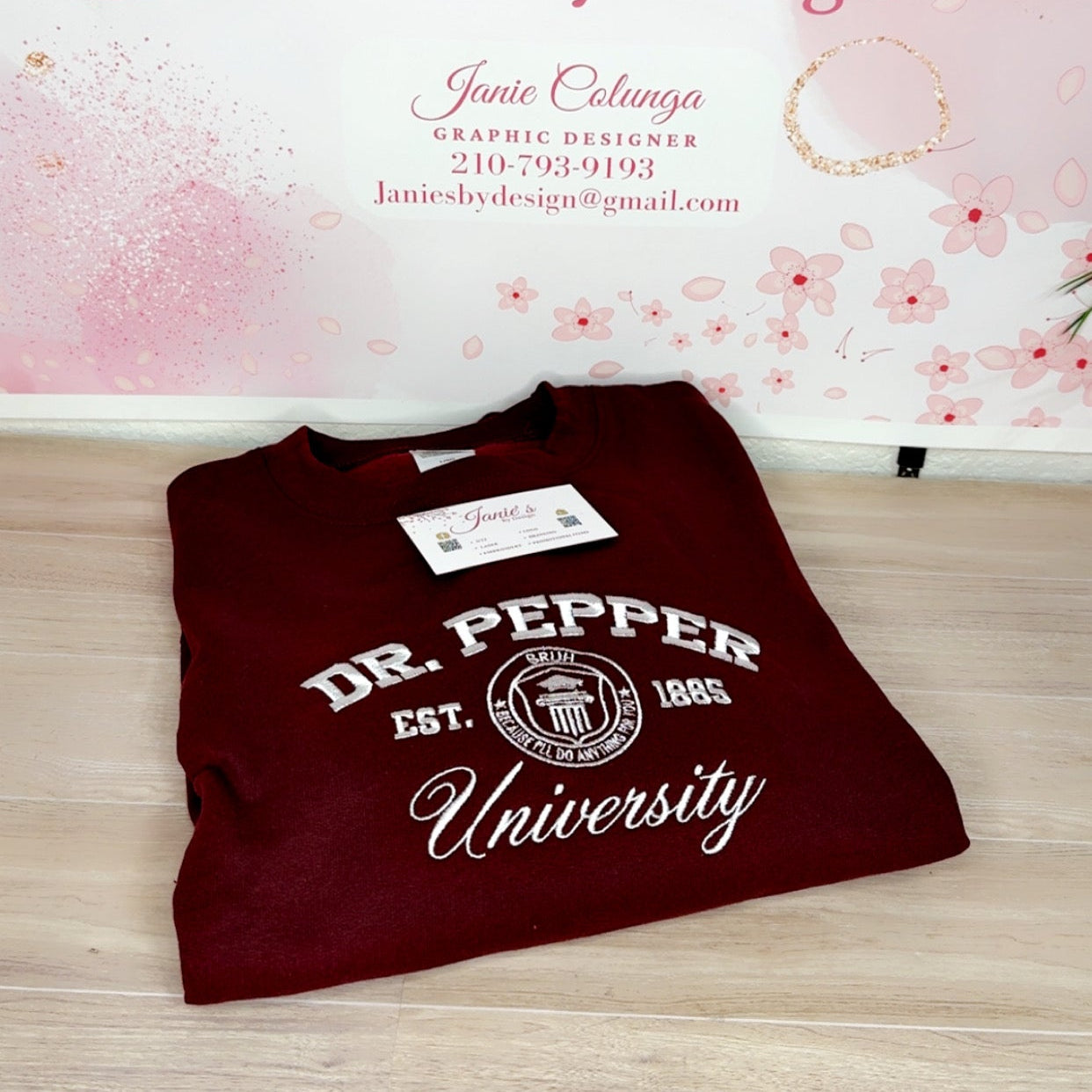 A folded maroon crewneck sweatshirt with 'DR. PEPPER University' printed in white on the front, displayed on a light wood floor in front of a pink floral backdrop with 'Janie's By Design' branding.