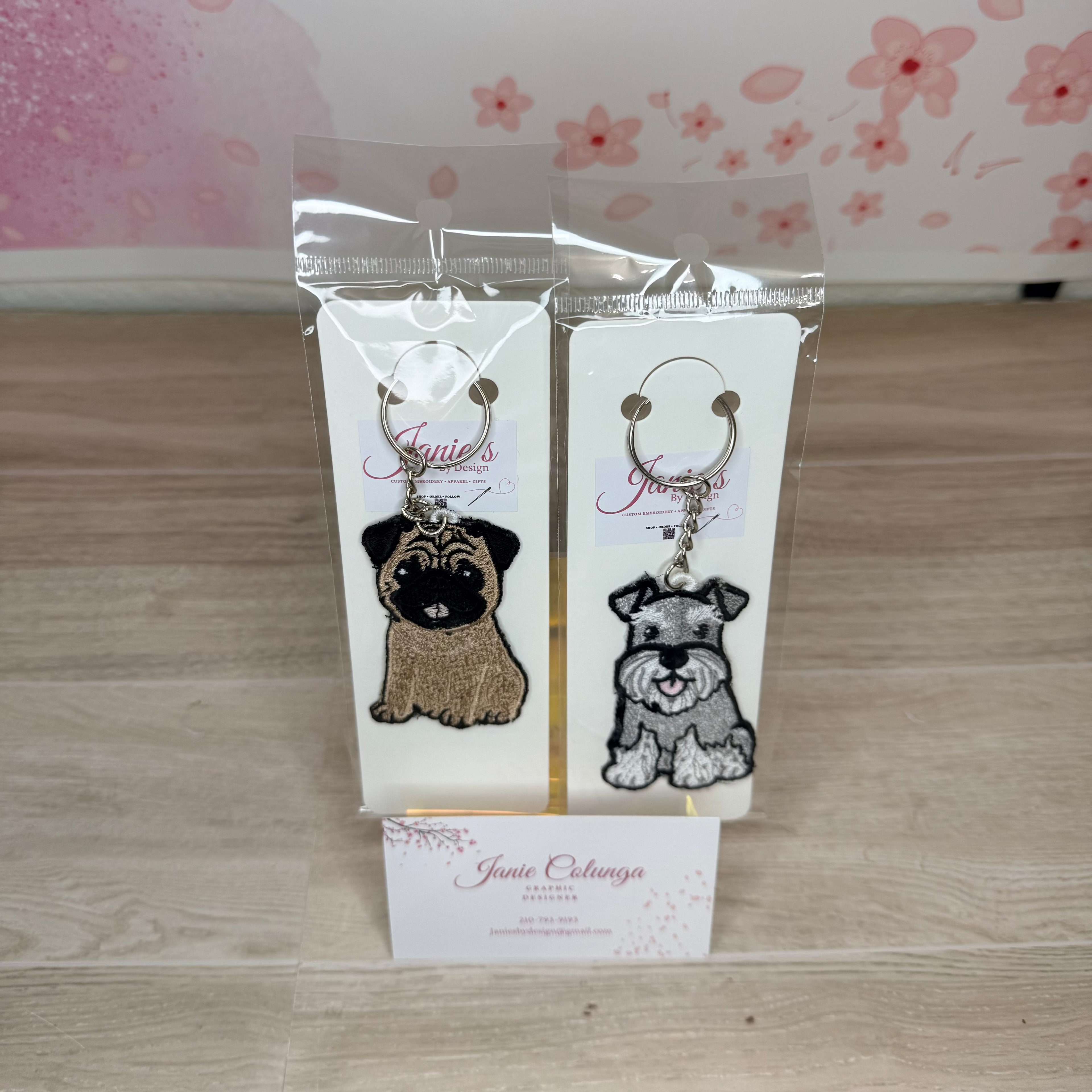 Two custom embroidered keychains featuring dog illustrations on white cards inside clear plastic packaging, displayed on a light wood surface with a pink floral background card.