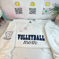 A white Gildan sweatshirt with the text 'Volleyball Mom' embroidered on the front, displayed on a flat surface with a QR code and social media tags in the background.
