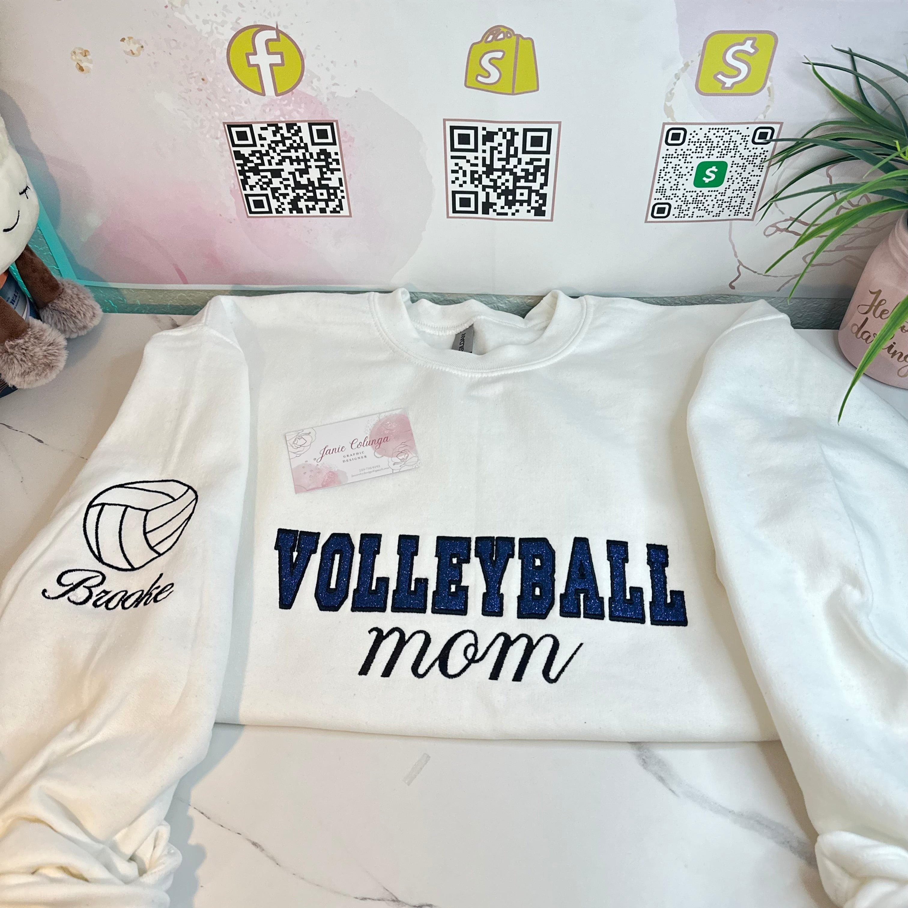 A white Gildan sweatshirt with the text 'Volleyball Mom' embroidered on the front, displayed on a flat surface with a QR code and social media tags in the background.