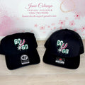 Two black caps with 'GO' and '47' logos on a wooden surface, branded 'Janie's By Design'.