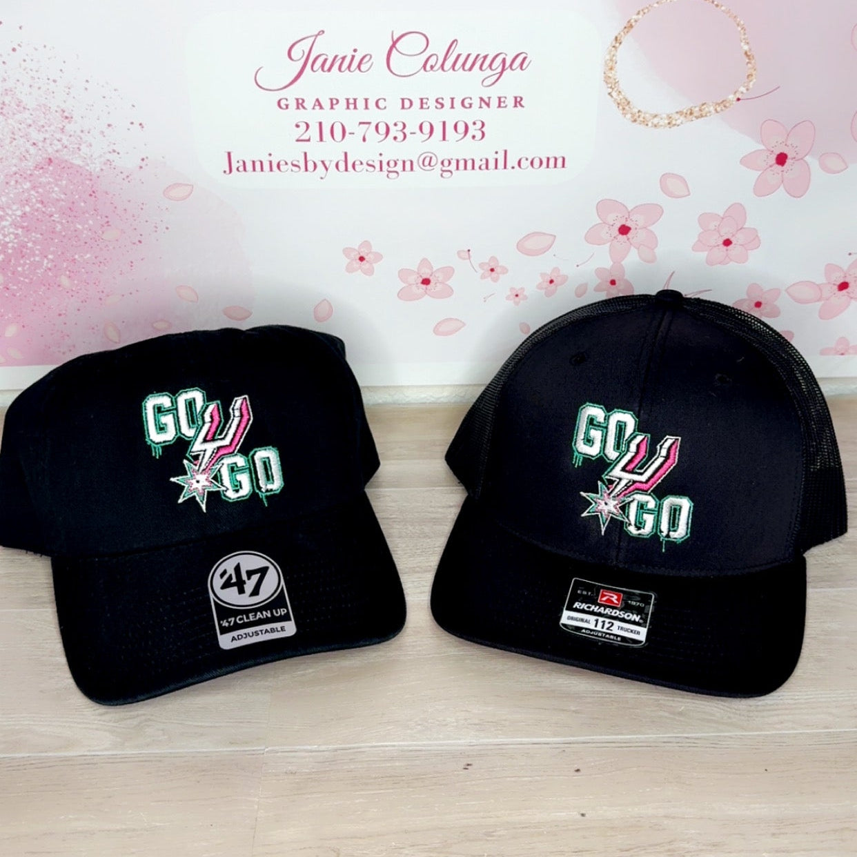 Two black caps with 'GO' and '47' logos on a wooden surface, branded 'Janie's By Design'.