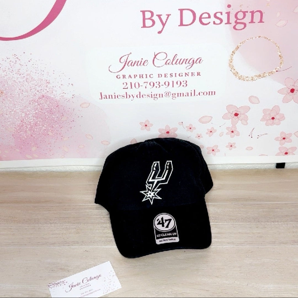 Black cap with a logo on a wooden surface in front of a 'Janie's by Design' branded background.