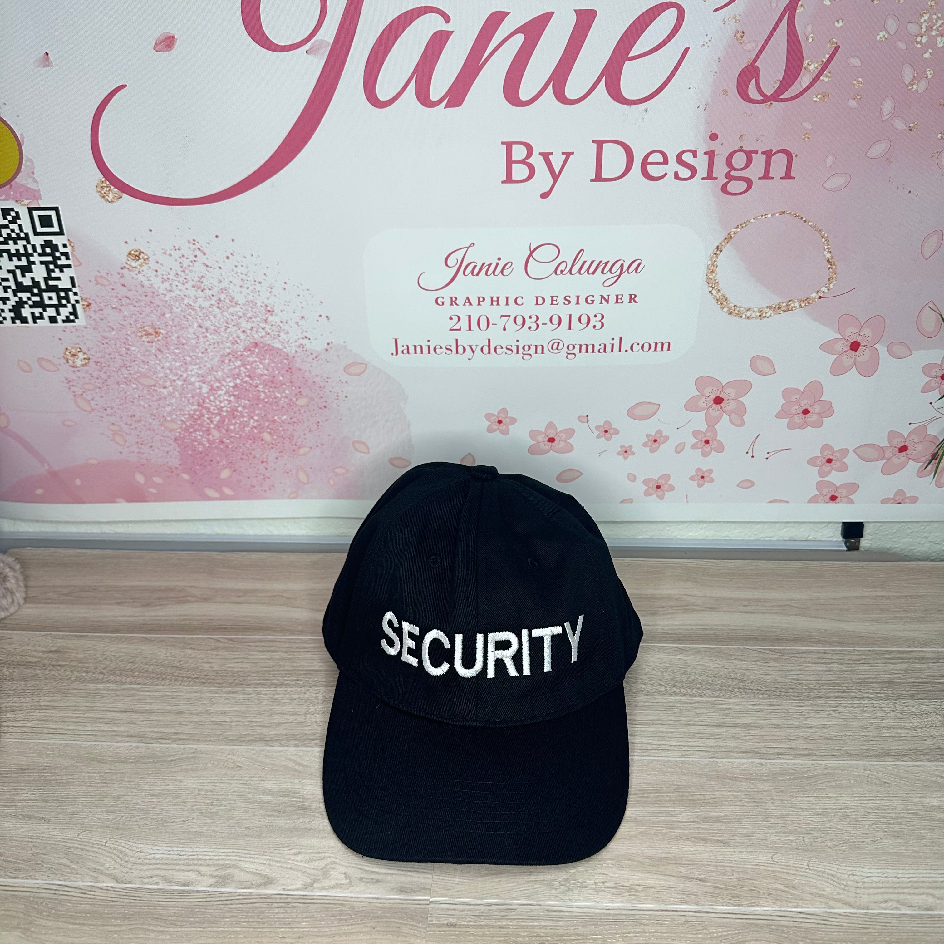 Black cap with 'SECURITY' text on a wooden surface in front of a decorative wall with 'Janie's By Design' branding.