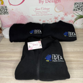 Black fleece jacket with IDEA Public Schools logo on a wooden surface, with 'Janie's by Design' branding in the background.