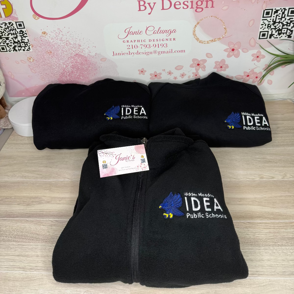 Black fleece jacket with IDEA Public Schools logo on a wooden surface, with 'Janie's by Design' branding in the background.