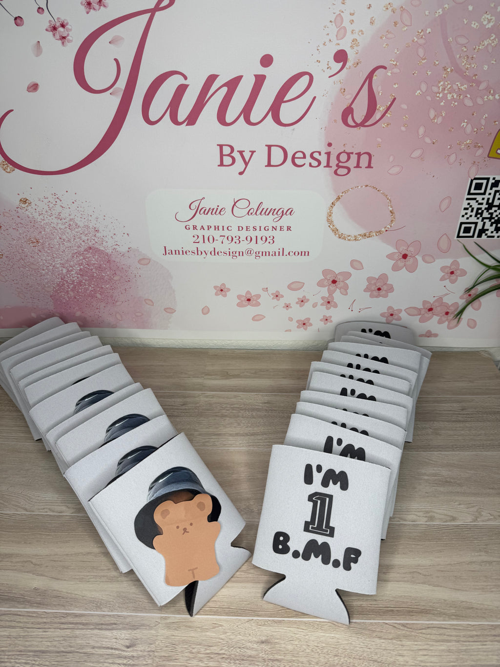A set of white fabric koozie bags with black printed text and graphics, arranged on a light wood floor in front of a pink floral backdrop. One bag shows a cartoon bear wearing a black hat, and another reads 'I'M 1 B.