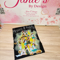 A black graphic t-shirt featuring a colorful print of cheerleaders and the text 'Heaven & Estella', displayed on a light wood floor in front of a pink branded backdrop that reads 'Janie's By Design'.