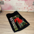 A black graphic t-shirt featuring Ronaldinho in a red soccer jersey with 'RONALDO' printed vertically in gold, laid flat on a light wood floor in front of a pink 'Janie's By Design' graphic wall.