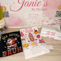 A styled product display featuring three graphic t-shirts from Janie's By Design on a light wood surface against a pink cherry blossom backdrop. The shirts include a black graphic with 'SIX SEVEN BRUH' and a Santa Claus illustration, a white graphic with 'IT'S THE SEASON' and a religious illustration, and a stack of folded white shirts with pink text.