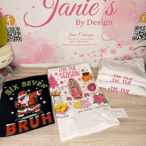 A styled product display featuring three graphic t-shirts from Janie's By Design on a light wood surface against a pink cherry blossom backdrop. The shirts include a black graphic with 'SIX SEVEN BRUH' and a Santa Claus illustration, a white graphic with 'IT'S THE SEASON' and a religious illustration, and a stack of folded white shirts with pink text.