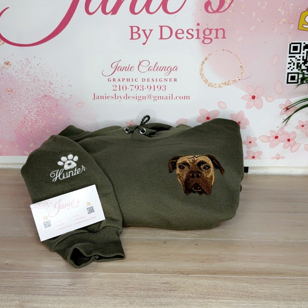 An olive green hoodie with a custom embroidered pug dog face on the chest and a matching embroidered sleeve cuff, displayed on a light wood floor in front of a pink 'Janie's By Design' graphic backdrop.