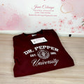 A folded maroon crewneck sweatshirt with 'DR. PEPPER University' printed in white on the front, displayed on a light wood floor in front of a pink floral backdrop with 'Janie's By Design' branding.