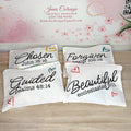 Four white decorative pillows with black inspirational Bible verse quotes are arranged on a light wood surface in front of a pink floral banner that reads 'Janie's By Design'.