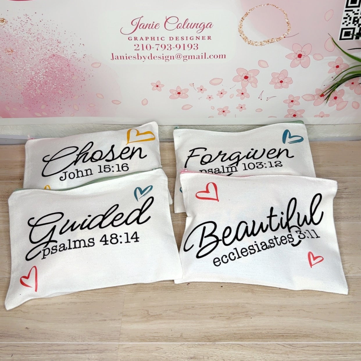 Four white decorative pillows with black inspirational Bible verse quotes are arranged on a light wood surface in front of a pink floral banner that reads 'Janie's By Design'.