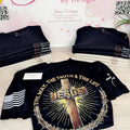 A black graphic t-shirt displayed on a table in front of a branded backdrop that reads 'Janie's By Design.' The shirt features a large front print with a cross, the word 'JESUS,' rays of light, and a crown of thorns, plus smaller printed graphics on the sleeves.