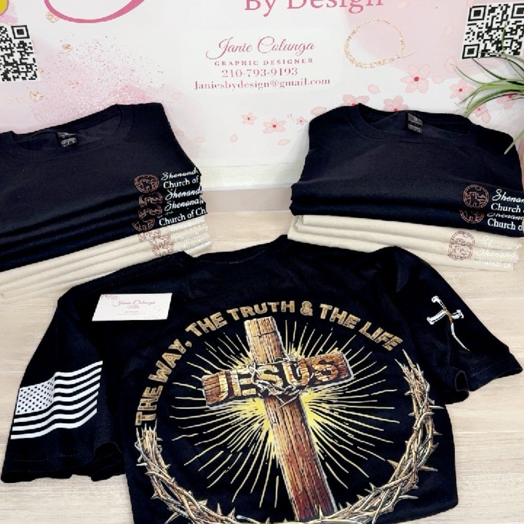 A black graphic t-shirt displayed on a table in front of a branded backdrop that reads 'Janie's By Design.' The shirt features a large front print with a cross, the word 'JESUS,' rays of light, and a crown of thorns, plus smaller printed graphics on the sleeves.