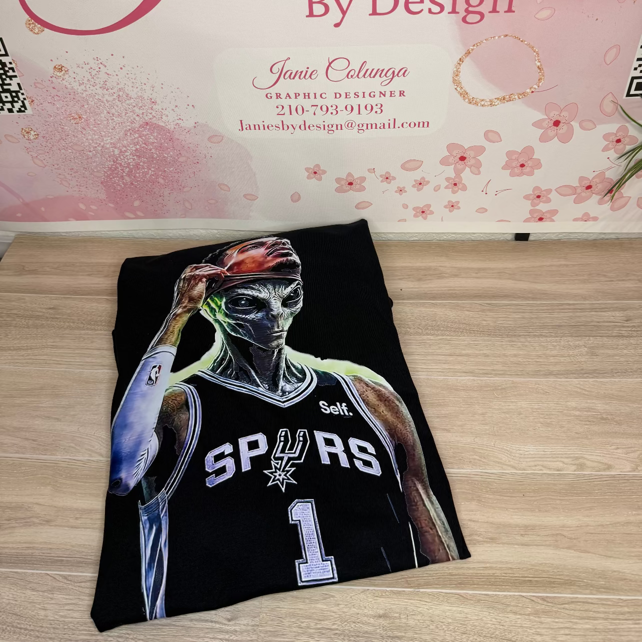 A black custom graphic t-shirt featuring a stylized illustration of a basketball player in a Spurs jersey, displayed on a light wood floor in front of a pink floral 'Janie's By Design' backdrop.