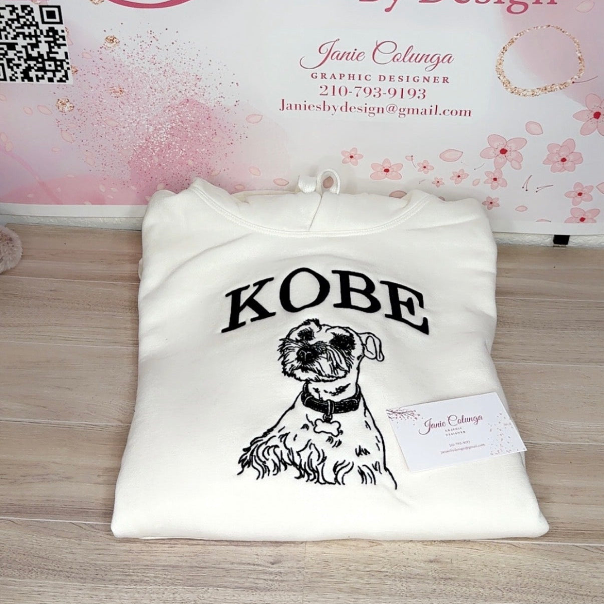 A folded white hoodie with a black graphic print of a dog and the word 'KOBE' is displayed on a light wood surface in front of a pink branded backdrop reading 'Janie's By Design'.