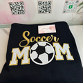 A folded black sweatshirt featuring a large embroidered design that reads 'Soccer MOM' in gold and white, with a soccer ball motif in the center. A small pink tag is attached near the top.