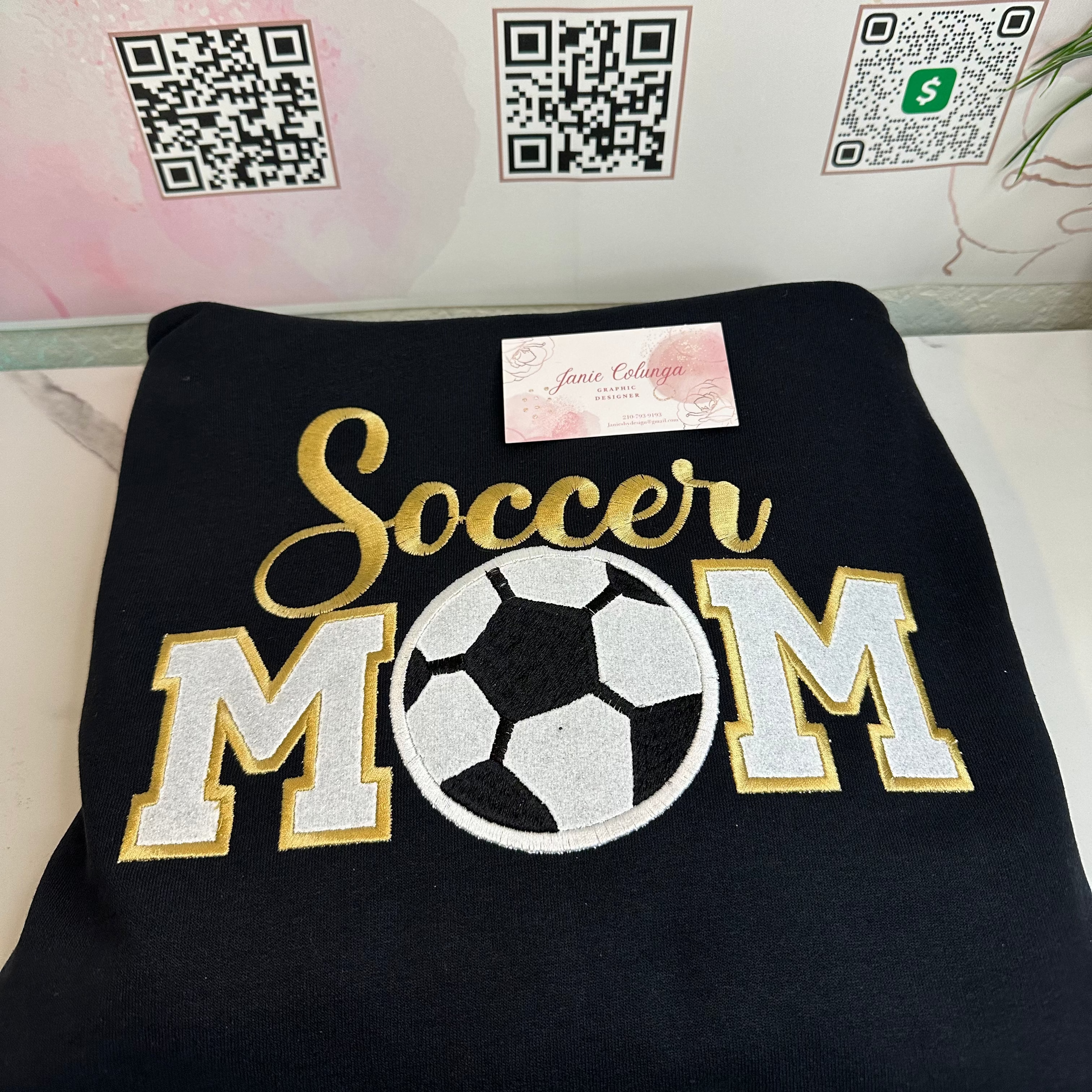 A folded black sweatshirt featuring a large embroidered design that reads 'Soccer MOM' in gold and white, with a soccer ball motif in the center. A small pink tag is attached near the top.
