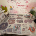 A display of various embroidered patches and appliques on a table, with a prominent sign reading 'Janie's By Design' in the background. The patches feature sports team designs including the Dallas Cowboys, Houston Rockets, San Antonio Spurs, and others in pink, silver, and blue thread.