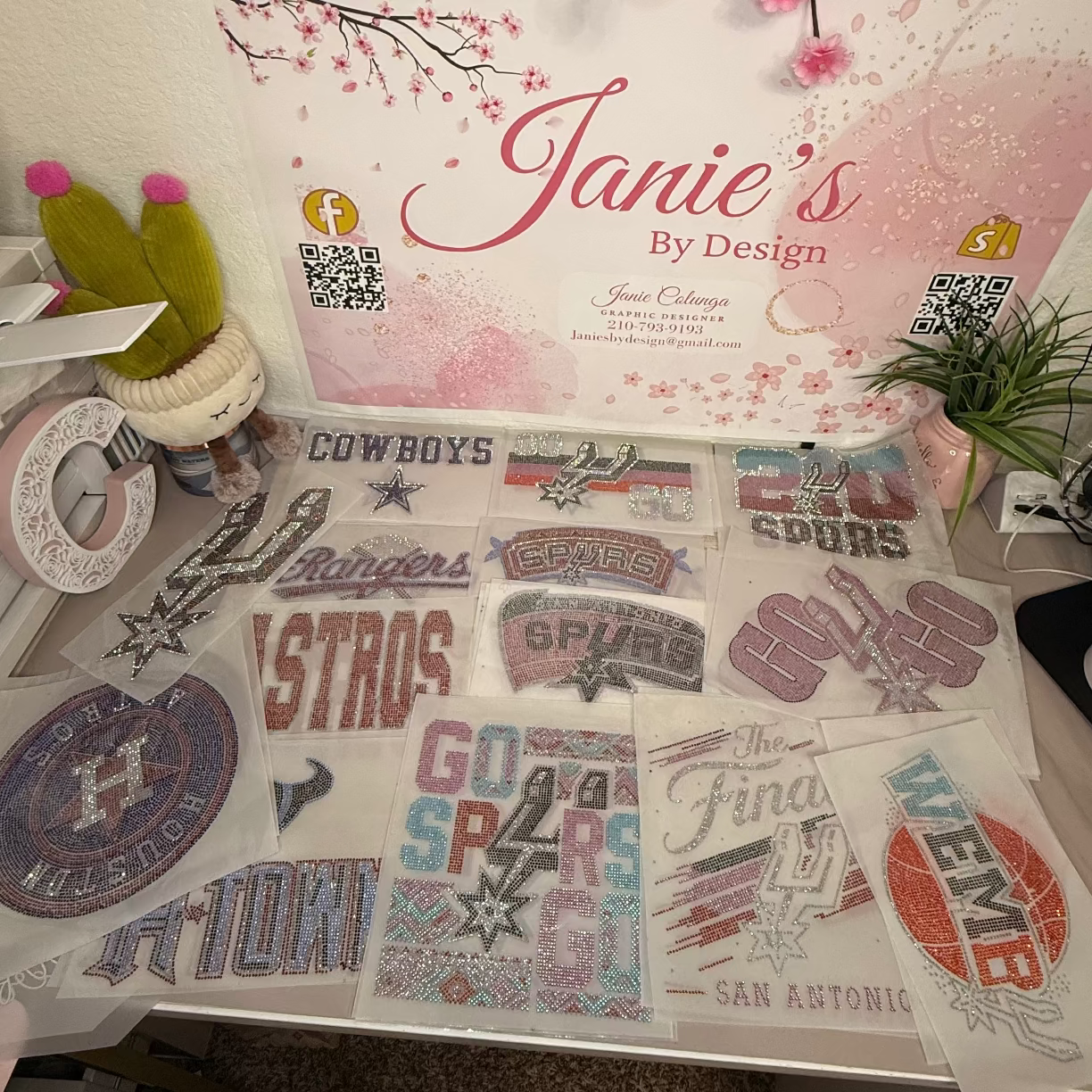 A display of various embroidered patches and appliques on a table, with a prominent sign reading 'Janie's By Design' in the background. The patches feature sports team designs including the Dallas Cowboys, Houston Rockets, San Antonio Spurs, and others in pink, silver, and blue thread.
