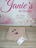 A folded beige knit blanket with pink and black embroidered text reading 'Such a Big Mother in such a little Girl' lies on a light wood floor in front of a branded wall sign that says 'Janie's By Design'. A small branded card is placed in front of the blanket.