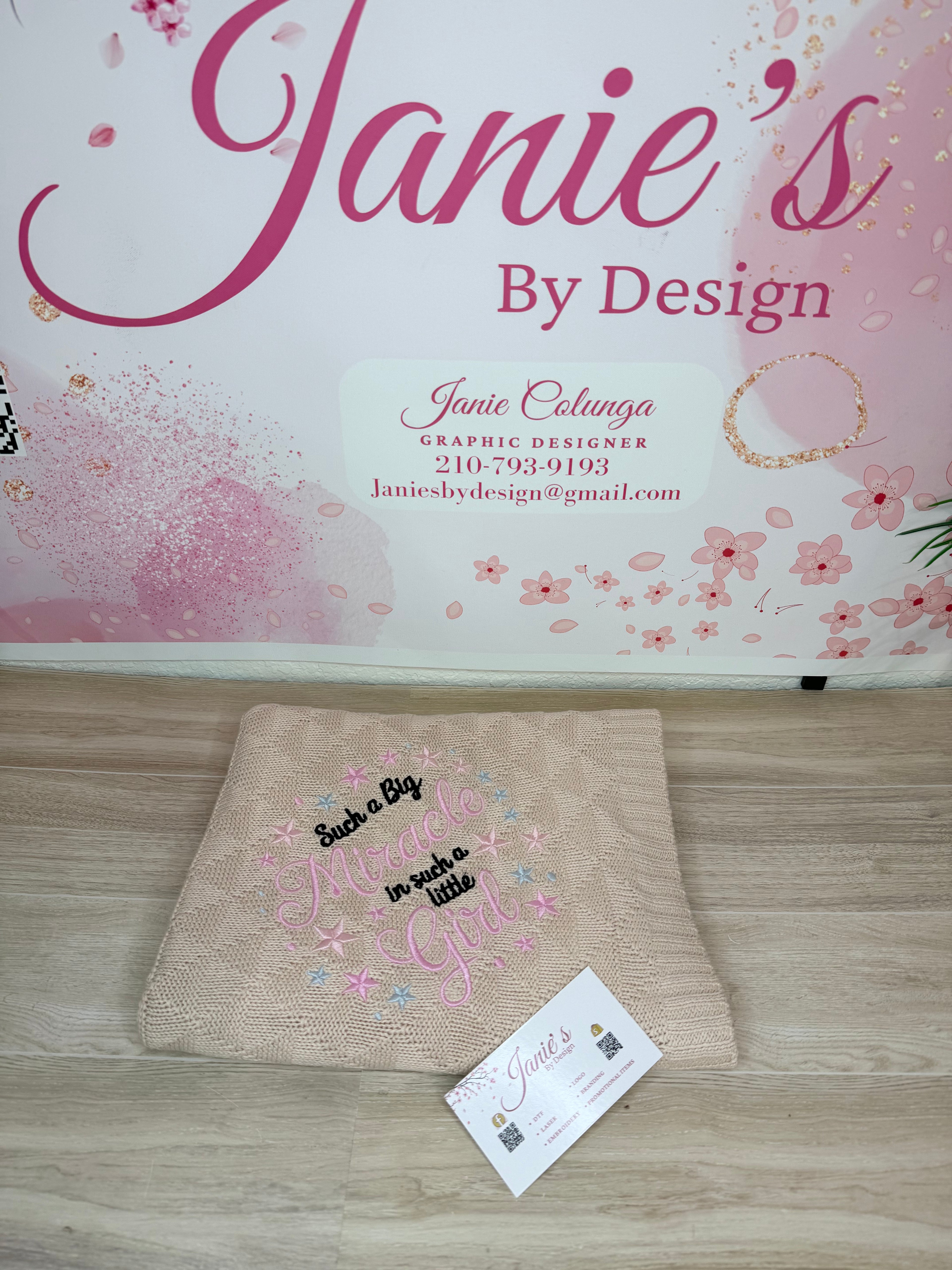 A folded beige knit blanket with pink and black embroidered text reading 'Such a Big Mother in such a little Girl' lies on a light wood floor in front of a branded wall sign that says 'Janie's By Design'. A small branded card is placed in front of the blanket.