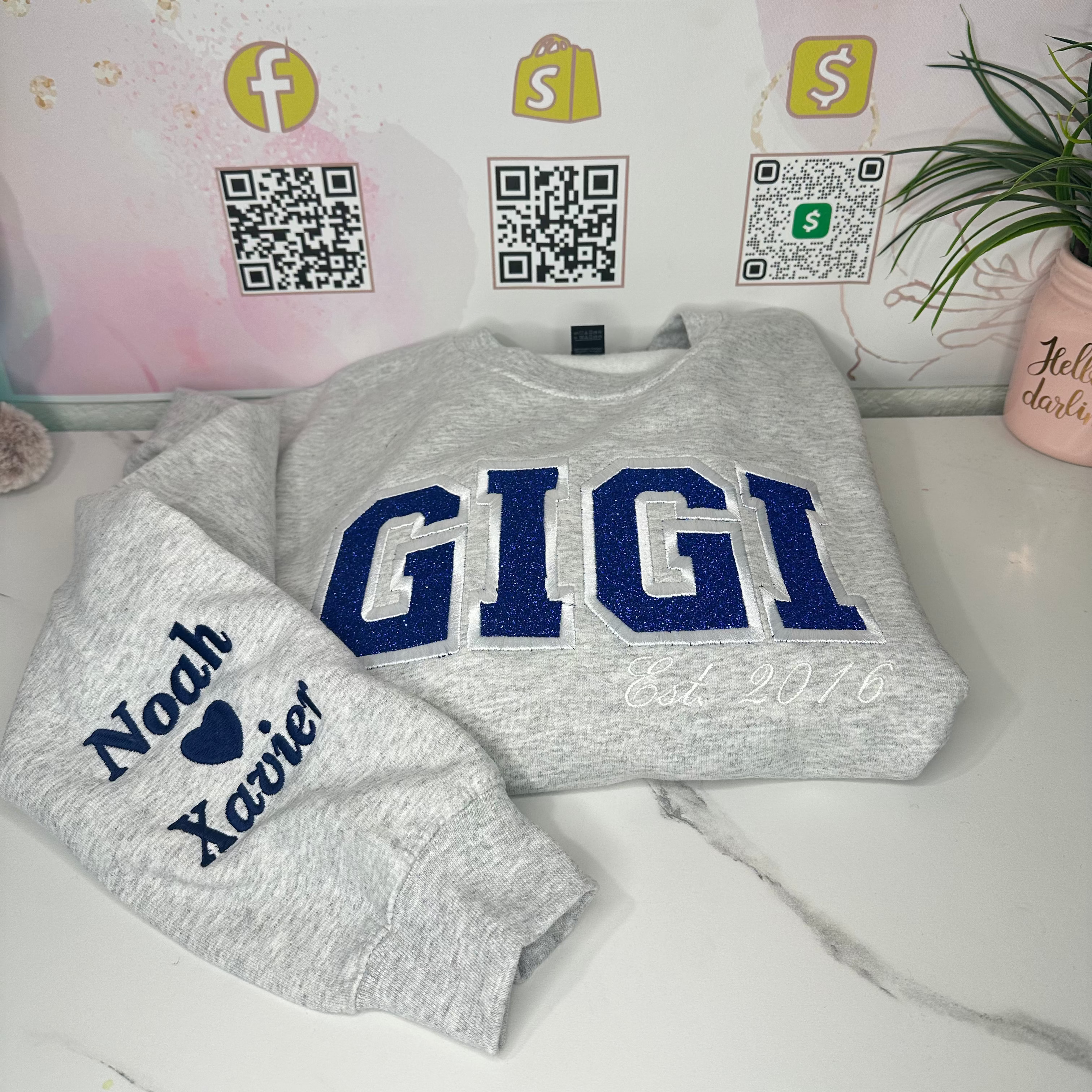 A light heather gray crewneck sweatshirt laid flat on a white marble surface, with large navy blue 'GIGI' lettering across the chest and a matching sleeve reading 'Noah x Xavier' in navy. The background shows a branded pink sign reading 'Janie's By Design' with QR codes and a small plant.