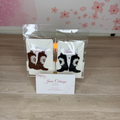 Two pairs of embroidered cowboy boot earrings displayed in clear plastic packaging on a light wood floor, with a business card in front and a pink floral wall in the background.