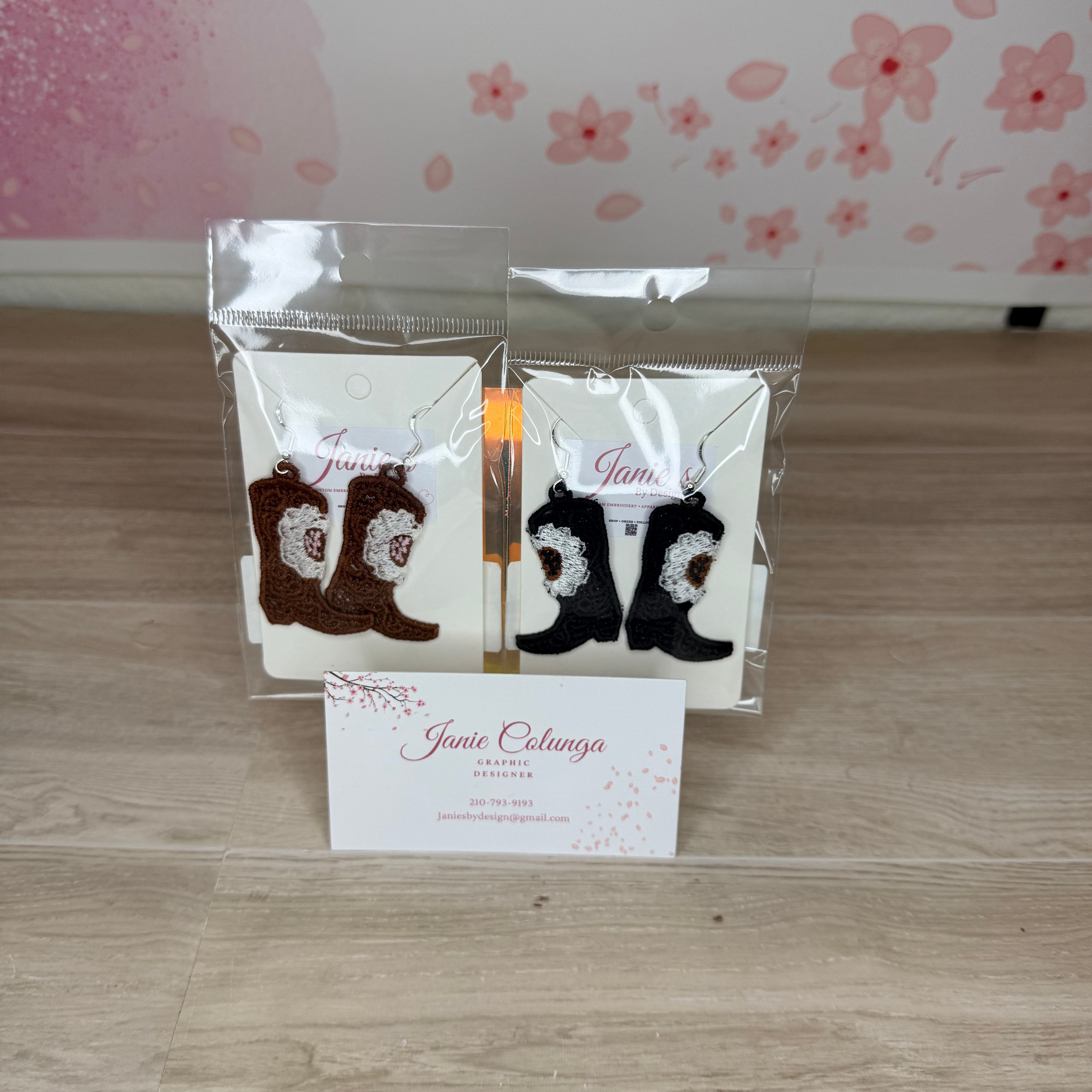 Two pairs of embroidered cowboy boot earrings displayed in clear plastic packaging on a light wood floor, with a business card in front and a pink floral wall in the background.