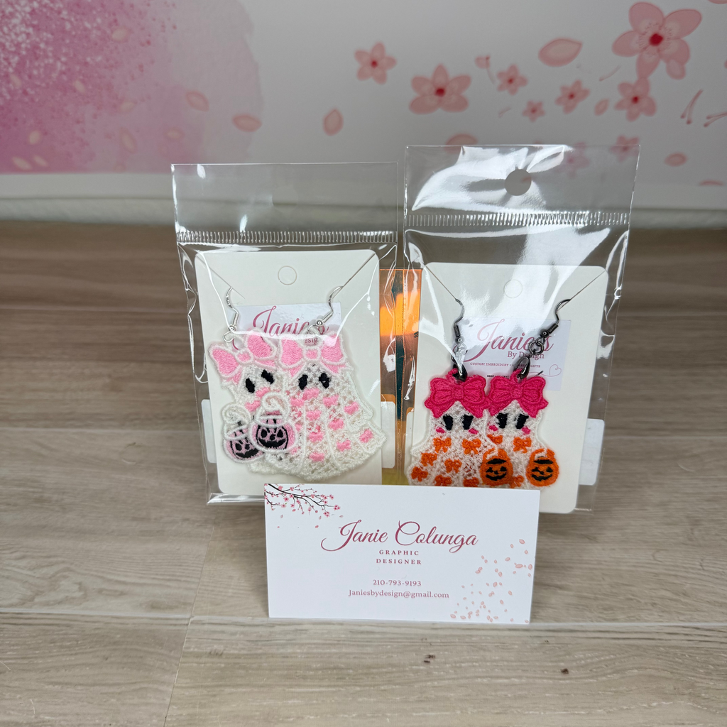 Two small packaged craft or accessory items displayed on a light wood surface, with a branded business card in front. The items are in clear plastic bags and appear to be white fabric or paper pieces decorated with pink and orange Halloween-themed designs, including bows, pumpkins, and black silhouettes.