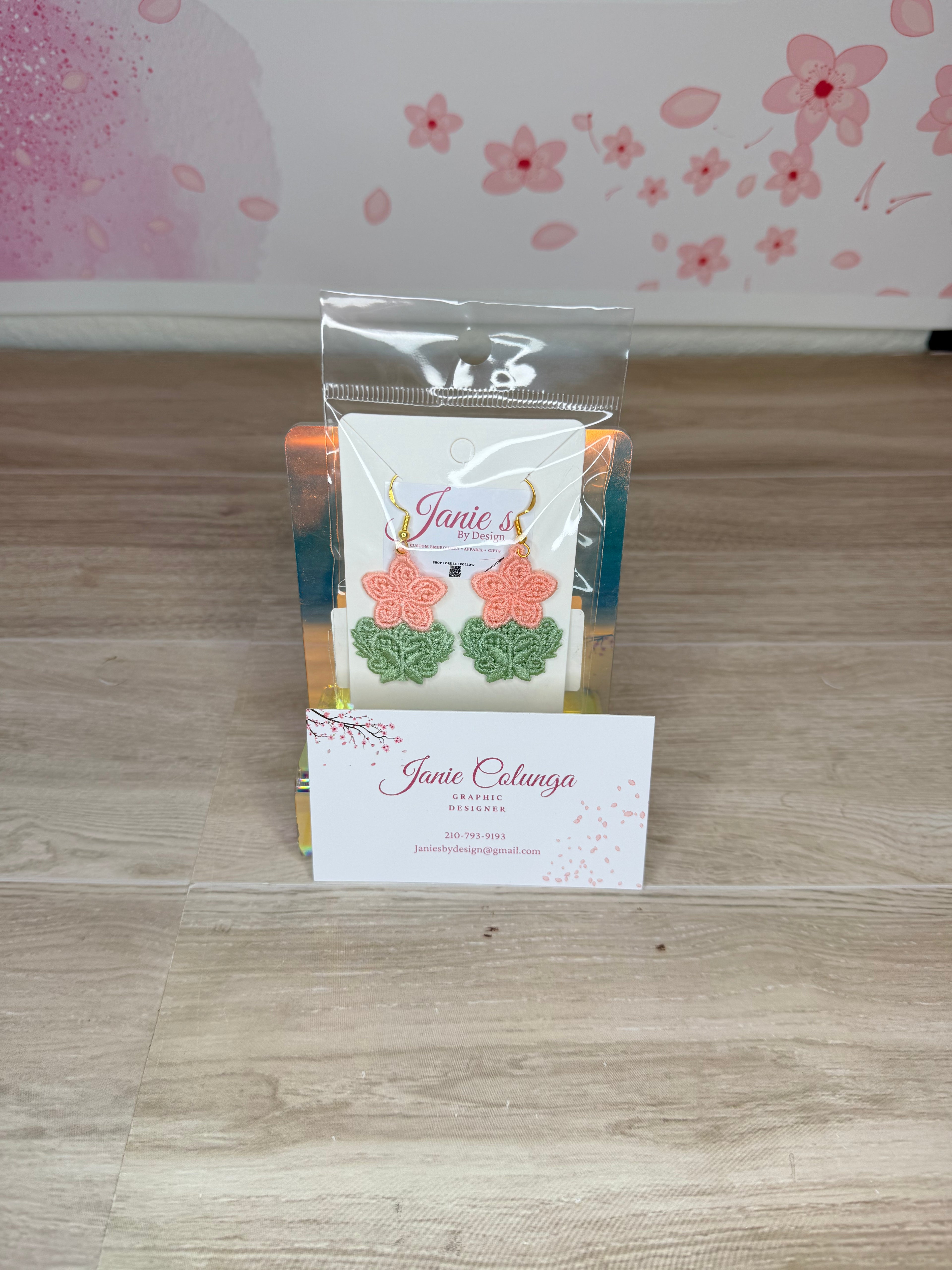 A pair of handmade coral-pink and green crochet earrings displayed on a white card inside a clear plastic bag, set on a light wood floor in front of a pink floral wall.