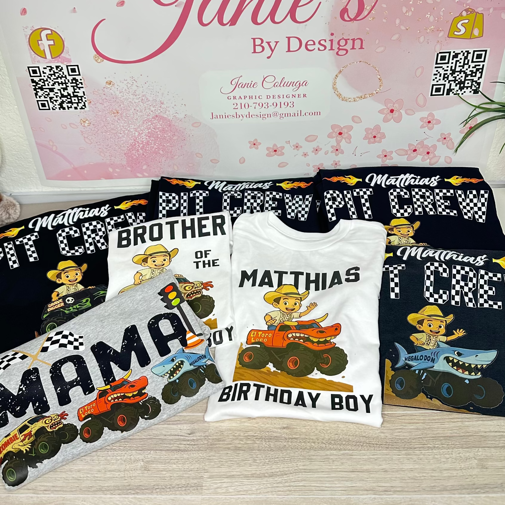 A set of custom printed t-shirts and a matching sweatshirt displayed on a light wood floor in front of a pink branded sign that reads 'Janie's By Design'. The shirts feature custom text like 'MATTHIAS BROTHER OF THE MATTHIAS BIRTHDAY BOY' and cartoon monster truck graphics on black, white, and gray shirts.