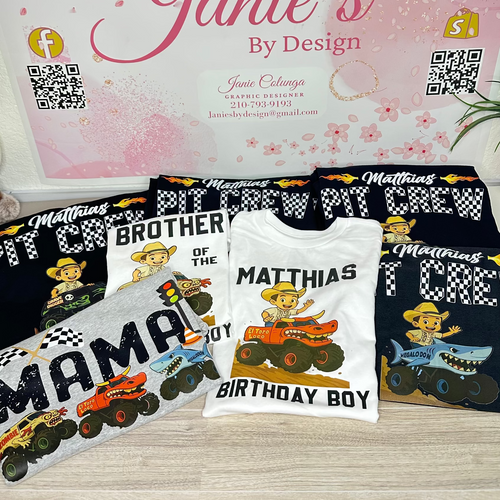 A set of custom printed t-shirts and a matching sweatshirt displayed on a light wood floor in front of a pink branded sign that reads 'Janie's By Design'. The shirts feature custom text like 'MATTHIAS BROTHER OF THE MATTHIAS BIRTHDAY BOY' and cartoon monster truck graphics on black, white, and gray shirts.