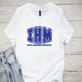 A white t-shirt with the text 'IHM' and 'Iowa Hidden Meow' printed in blue on the front, displayed on a wooden surface with jeans to the side.
