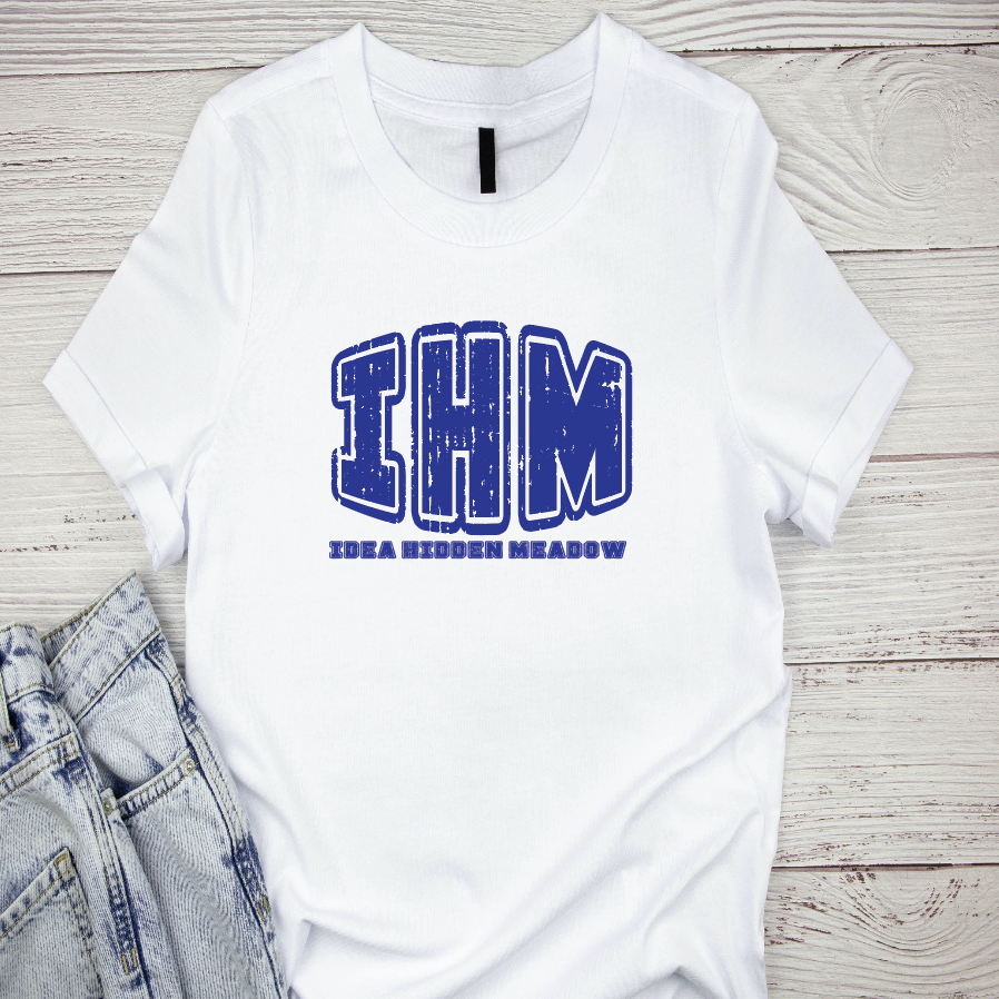 A white t-shirt with the text 'IHM' and 'Iowa Hidden Meow' printed in blue on the front, displayed on a wooden surface with jeans to the side.
