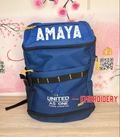 Blue backpack with 'AMAYA' branding on a wooden surface with floral decorations in the background.