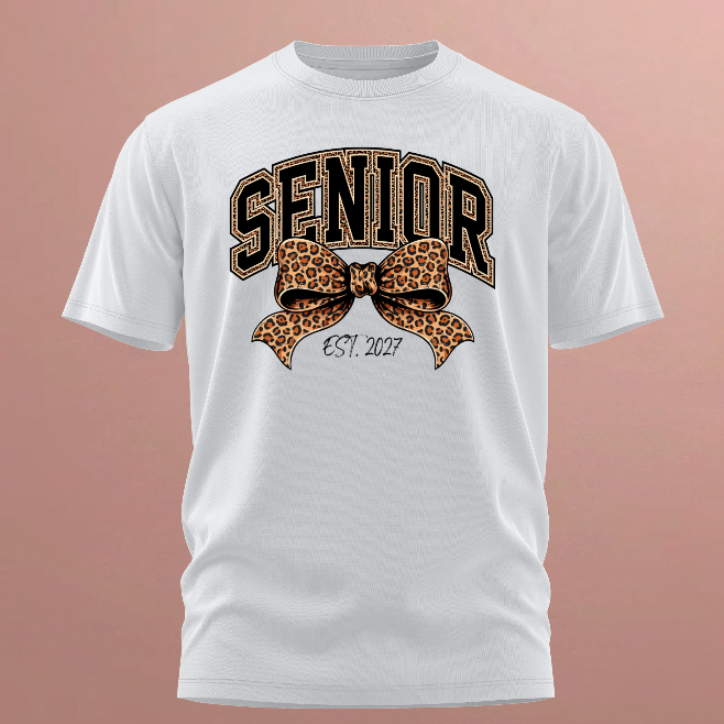 A white short-sleeve crewneck t-shirt with a graphic on the front featuring the word 'SENIOR' in arched brown letters with leopard print outlines, a matching leopard print bow, and 'EST. 2027' written below, shown against a solid dusty rose background.