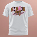 A white short-sleeve t-shirt with a 'Senior Class of 2027' graphic on the chest, featuring leopard print and pink details, displayed against a solid pinkish background.