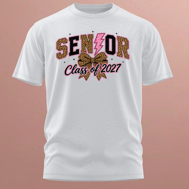 A white short-sleeve t-shirt with a 'Senior Class of 2027' graphic on the chest, featuring leopard print and pink details, displayed against a solid pinkish background.