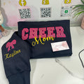 Navy sweatshirt with 'CHEER Mom' and 'Kaylinn' text, displayed on a white surface with a branded backdrop.