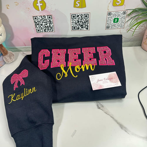 Navy sweatshirt with 'CHEER Mom' and 'Kaylinn' text, displayed on a white surface with a branded backdrop.